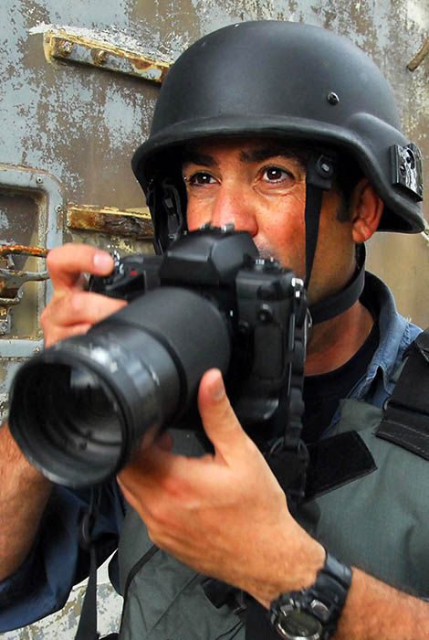 Photojournalist in protective gear takes photo in war zone.