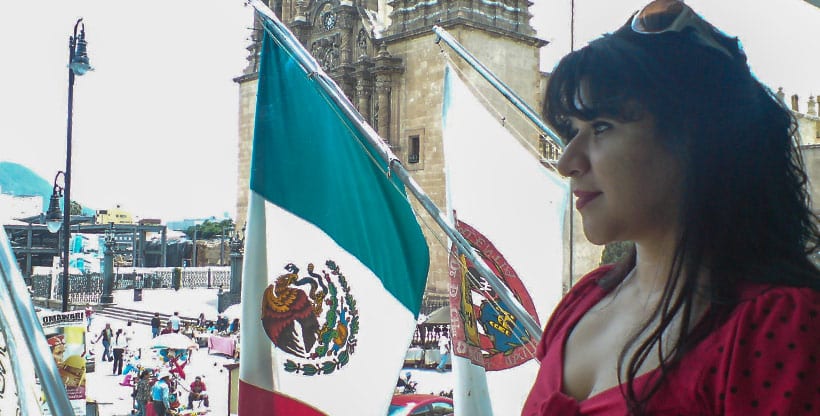 Expat woman on a balcony next to a Mexican flag, looking over a busy plaza with colonial architecture and street vendors.