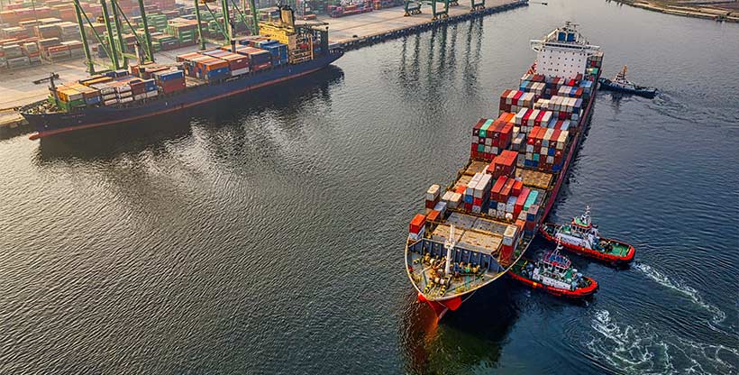 A cargo ship loaded with multicolored shipping containers is pushed to an unloading dock by tugboats.